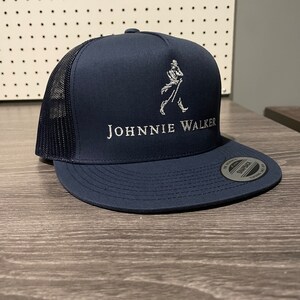 May include: A navy blue trucker hat with a black mesh back. The hat has a white embroidered Johnnie Walker logo with a walking man figure.