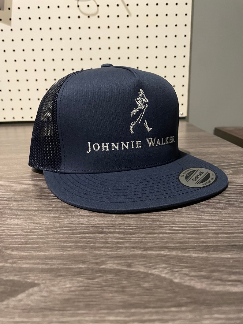 May include: A navy blue trucker hat with a black mesh back. The hat has a white embroidered Johnnie Walker logo with a walking man figure.