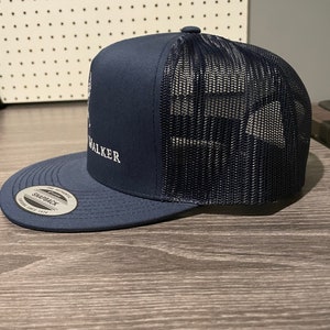 May include: A navy blue trucker hat with a black mesh back. The hat has a white embroidered logo that says "WALKER" on the front.