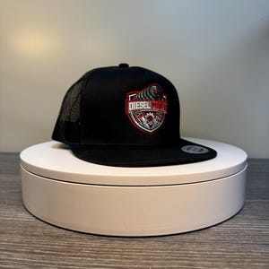 Pode incluir: Bon&eacute; de caminhoneiro preto com parte traseira em malha e aba reta. A frente do bon&eacute; apresenta um design bordado em vermelho, branco e cinza com as palavras "DIESEL POWER" em forma de escudo.