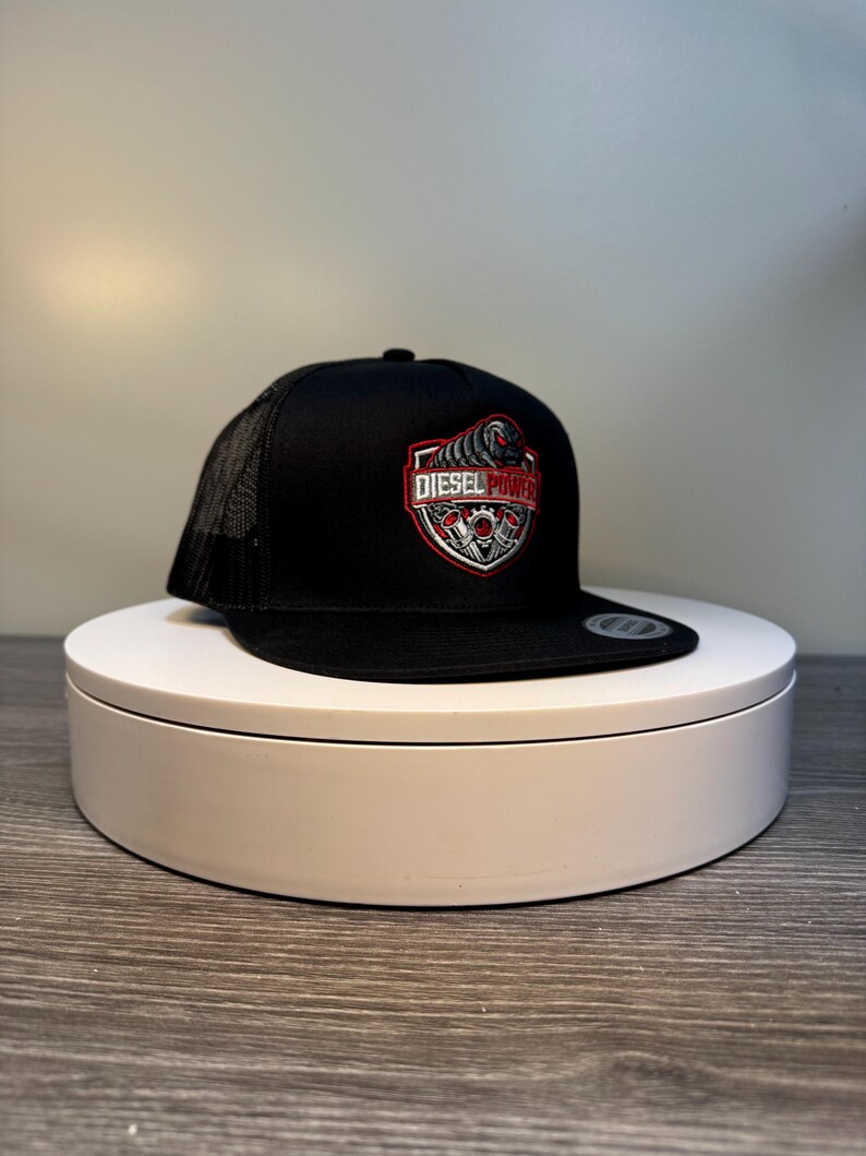 Pode incluir: Bon&eacute; de caminhoneiro preto com parte traseira em malha e aba reta. A frente do bon&eacute; apresenta um design bordado em vermelho, branco e cinza com as palavras "DIESEL POWER" em forma de escudo.