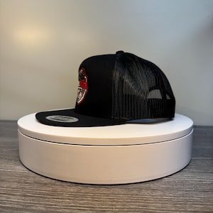 Pode incluir: Bon&eacute; de caminhoneiro preto com costas em malha e aba reta. A frente do bon&eacute; apresenta um emblema vermelho e branco. O bon&eacute; &eacute; exibido em uma plataforma girat&oacute;ria branca.