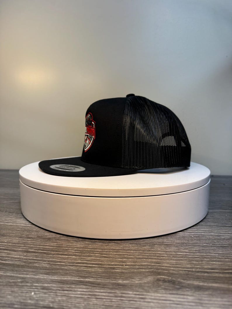 Pode incluir: Bon&eacute; de caminhoneiro preto com costas em malha e aba reta. A frente do bon&eacute; apresenta um emblema vermelho e branco. O bon&eacute; &eacute; exibido em uma plataforma girat&oacute;ria branca.