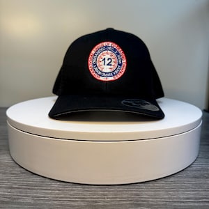 May include: Black baseball cap with a circular patch featuring the text "International Union of Operating Engineers" and the number "12". The patch has red, white, and blue accents. The hat is displayed on a white rotating stand.