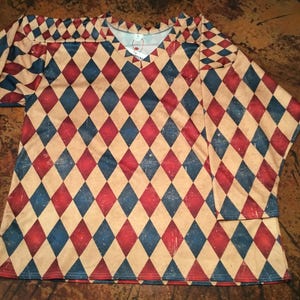 May include: A long-sleeved shirt with a V-neck, featuring a repeating diamond pattern in red, blue, and tan. The design has a distressed, vintage look. The shirt is made of a soft, lightweight material.