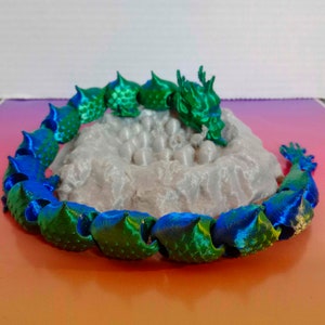 Puede incluir: Un dragón articulado impreso en 3D con un acabado degradado en verde y azul metálico, enroscado alrededor de un nido gris texturizado que contiene varios huevos pequeños, redondeados y de color gris claro. El dragón tiene escamas detalladas.