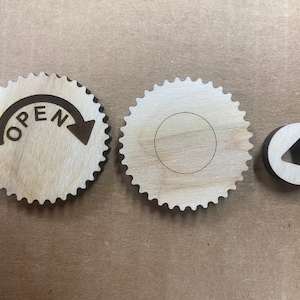 Può includere: Tre ingranaggi di legno di forma circolare. Il primo ingranaggio ha la scritta "OPEN" e una freccia che punta a destra. Il secondo ingranaggio ha un foro circolare al centro. Il terzo ingranaggio ha una forma triangolare tagliata al centro.