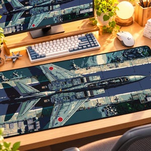 May include: A rectangular desk mat featuring a detailed top-down illustration of a grey fighter jet with red circular insignias on its wings. The jet is positioned over a stylized airfield runway graphic with dark blue and green accents.