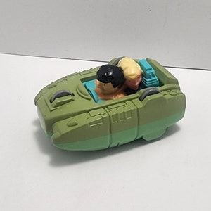 May include: A green plastic toy spaceship with two figures inside. The figures are a man with black hair and a woman with blonde hair.