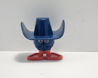 May include: A novelty toy figure with a blue cowboy hat, a blue face with a moustache, and red feet. The figure has two small, round eyes. The toy is designed to stand upright.