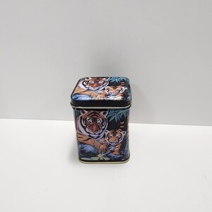 May include: A black square tin with a colorful image of a tiger and two cubs in a jungle setting.