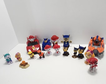 May include: A collection of Paw Patrol figurines, including Chase, Marshall, Skye, and Rubble. The figurines are in various poses and come with their vehicles. There is also a campfire and a small dog figurine.