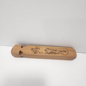 Peut inclure: Un sifflet en bois de couleur marron clair avec le texte "Capt Al's" et une image de dessin animé d'un train et d'un conducteur. Le sifflet est rectangulaire avec une ouverture.