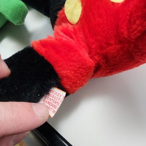 May include: Close-up of a plush Mickey Mouse glove. The glove is red with yellow circles and black accents. A tag with text is visible. The plush toy is a classic Disney character.