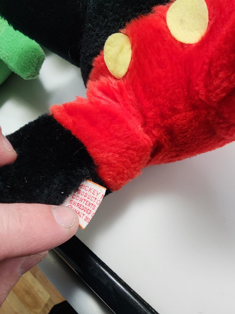 May include: Close-up of a plush Mickey Mouse glove. The glove is red with yellow circles and black accents. A tag with text is visible. The plush toy is a classic Disney character.