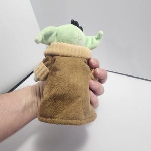 May include: A plush toy of a character with large green ears and a brown robe. The toy has a light green face and a tan collar. The character is from a popular science fiction franchise.