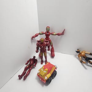 K&ouml;nnte beinhalten: Eine Sammlung von Superhelden-Actionfiguren und einem Spielzeugauto. Die Figuren umfassen Iron Man in verschiedenen Posen und eine Wolverine-Figur. Das Spielzeugauto ist rot und goldfarben mit schwarzen R&auml;dern. Die Figuren befinden sich auf einer wei&szlig;en Oberfl&auml;che.