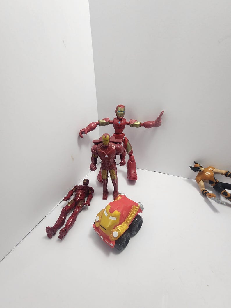 K&ouml;nnte beinhalten: Eine Sammlung von Superhelden-Actionfiguren und einem Spielzeugauto. Die Figuren umfassen Iron Man in verschiedenen Posen und eine Wolverine-Figur. Das Spielzeugauto ist rot und goldfarben mit schwarzen R&auml;dern. Die Figuren befinden sich auf einer wei&szlig;en Oberfl&auml;che.