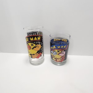 May include: Two clear glass drinking glasses with Pac-Man graphics. The glasses feature a black and yellow Pac-Man character with red and blue ghosts. The text "Pac-Man" is printed on the front of each glass.