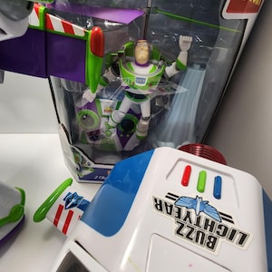 May include: A green and purple toy helicopter with a Buzz Lightyear figure in the cockpit. The helicopter is in a clear plastic box with a white background. The box has the words "Toy Story" printed on it. The helicopter has a red and white striped tail and a green propeller. The figure is wearing a green and white spacesuit with a purple helmet. The figure has a red and white striped belt and a green and white spacesuit.