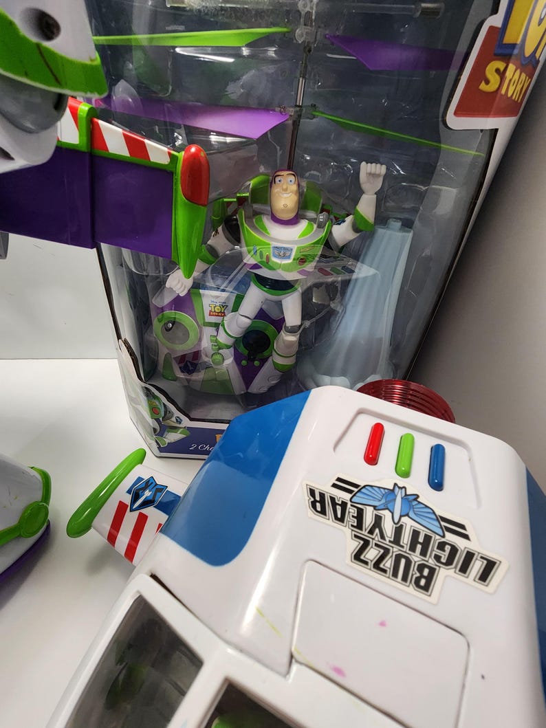 May include: A green and purple toy helicopter with a Buzz Lightyear figure in the cockpit. The helicopter is in a clear plastic box with a white background. The box has the words "Toy Story" printed on it. The helicopter has a red and white striped tail and a green propeller. The figure is wearing a green and white spacesuit with a purple helmet. The figure has a red and white striped belt and a green and white spacesuit.