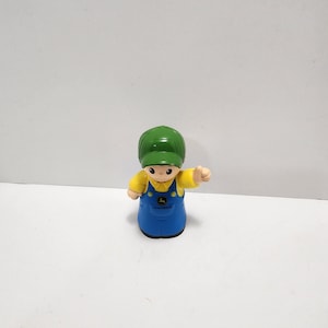May include: A green and yellow plastic toy figure of a person wearing a green hat and blue overalls with the John Deere logo. The figure is giving a thumbs up.