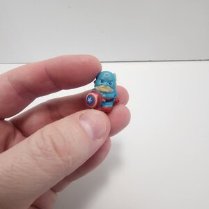 May include: A small plastic figurine of Captain America, wearing his iconic blue and red costume, holding his shield. The figurine is about 1 inch tall.