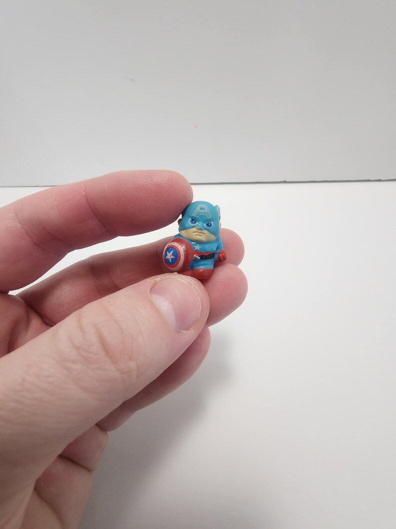 May include: A small plastic figurine of Captain America, wearing his iconic blue and red costume, holding his shield. The figurine is about 1 inch tall.