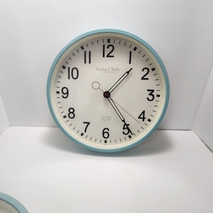 May include: A round wall clock with a light blue frame and a white face. The clock features black numerals and hands, with the brand name "Sterling & Noble" printed at the top. The clock measures approximately 10 inches in diameter.