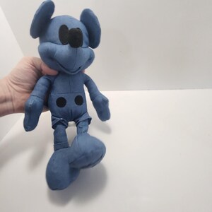 Puede incluir: Un peluche azul de un rat&oacute;n de dibujos animados con orejas grandes, ojos negros y una nariz negra. El juguete tiene un dise&ntilde;o cosido y dos detalles de botones negros en la parte delantera. El juguete se sostiene en una mano sobre un fondo blanco.
