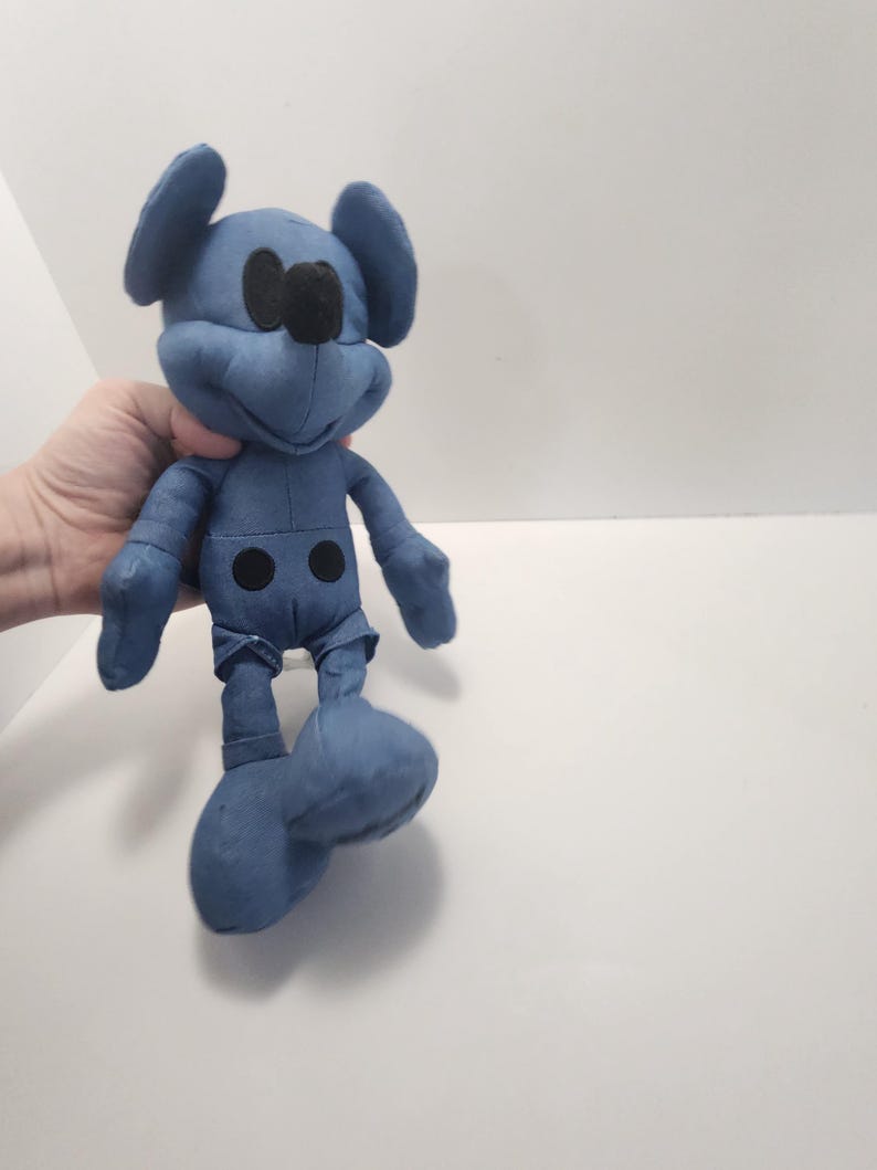 Puede incluir: Un peluche azul de un rat&oacute;n de dibujos animados con orejas grandes, ojos negros y una nariz negra. El juguete tiene un dise&ntilde;o cosido y dos detalles de botones negros en la parte delantera. El juguete se sostiene en una mano sobre un fondo blanco.