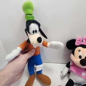 May include: A plush toy of Goofy wearing a green hat and orange shirt with blue trousers.  A plush toy of Minnie Mouse wearing a pink bow and pink shoes is also in the image.