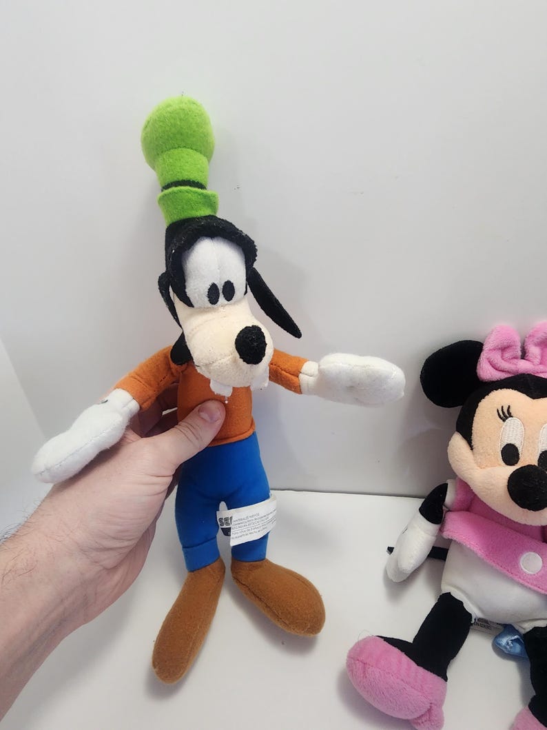 May include: A plush toy of Goofy wearing a green hat and orange shirt with blue trousers.  A plush toy of Minnie Mouse wearing a pink bow and pink shoes is also in the image.