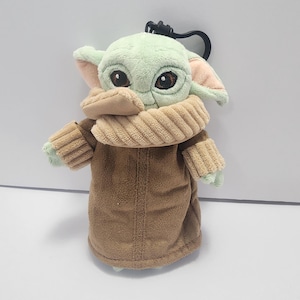 May include: A plush keychain featuring a character with large eyes, green skin, and large ears. The character wears a brown coat and a tan scarf. The keychain has a black clasp.