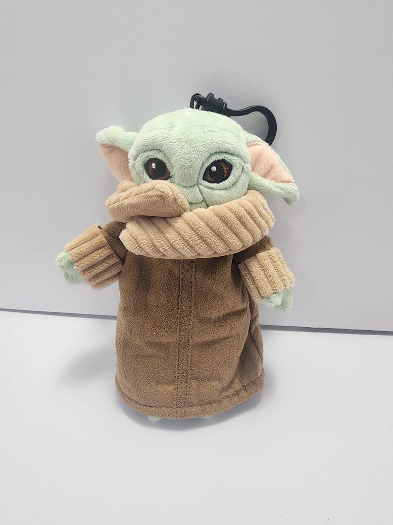 May include: A plush keychain featuring a character with large eyes, green skin, and large ears. The character wears a brown coat and a tan scarf. The keychain has a black clasp.
