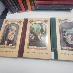 May include: A collection of hardcover books from the "A Series of Unfortunate Events" series by Lemony Snicket. The books have colorful spines and illustrations on the covers. Titles include "The Bad Beginning" and "The Reptile Room."