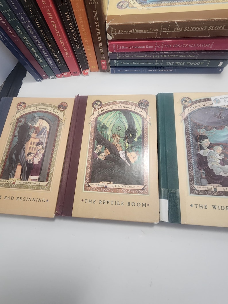 May include: A collection of hardcover books from the "A Series of Unfortunate Events" series by Lemony Snicket. The books have colorful spines and illustrations on the covers. Titles include "The Bad Beginning" and "The Reptile Room."