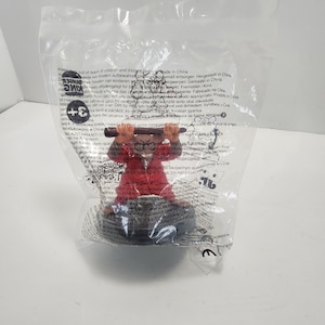 May include: A Burger King promotional figurine of a character wearing a red outfit and holding a brown stick, sealed inside a clear plastic bag. The packaging features printed safety warnings and the Burger King logo in black text on the front.
