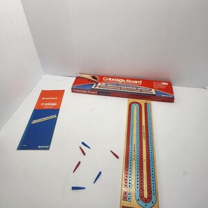 May include: A cribbage board game set with a wooden board, red and blue pegs, and a box. The box is red with the words "Cribbage Board" in white. A blue and orange instruction card is also included.