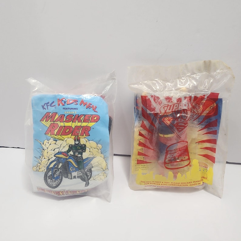 May include: Two sealed KFC Kids Meal toys. One features "Masked Rider" with a motorcycle graphic. The other features "Superman" with a burger graphic. Both are in clear plastic packaging.