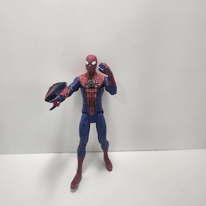 May include: A Spider-Man action figure in a dynamic pose, featuring a red and blue costume with web detailing. The figure is holding a black and blue object in its left hand. The figure is standing on a white surface.