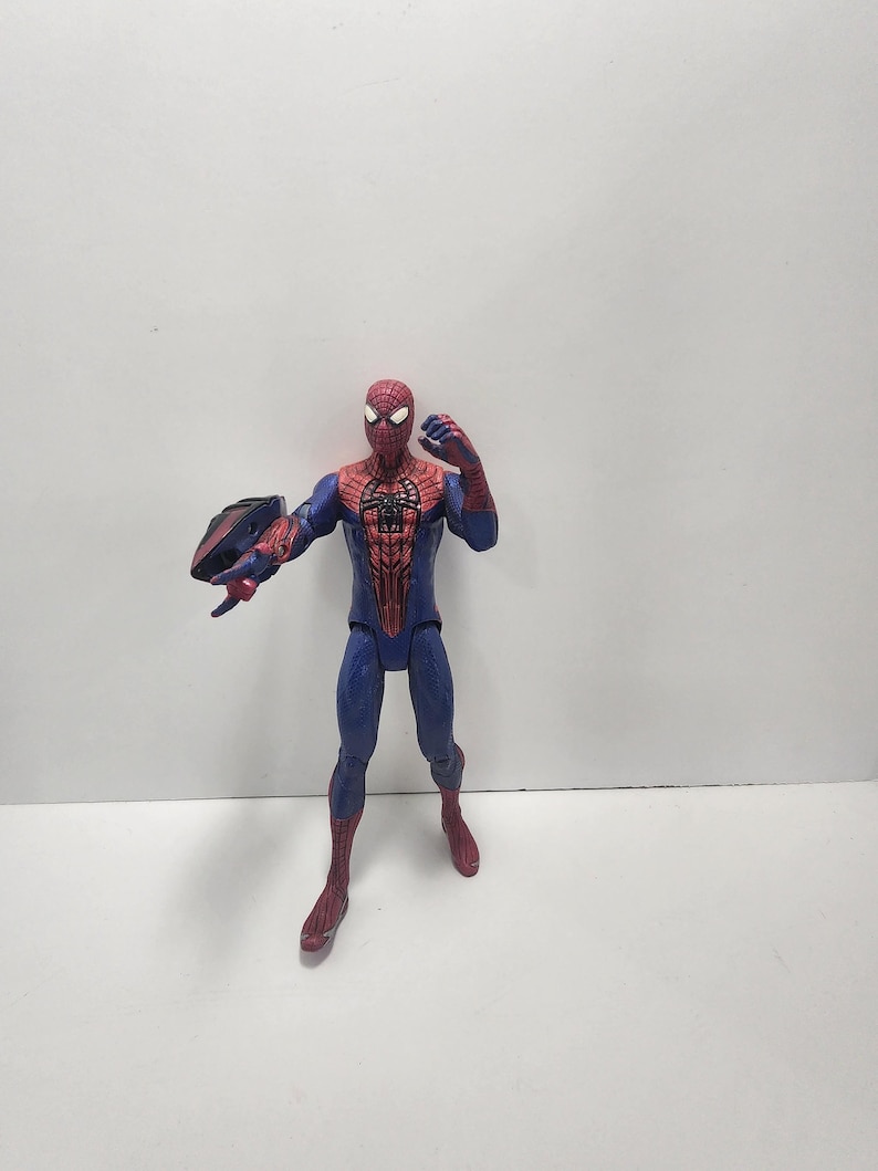 May include: A Spider-Man action figure in a dynamic pose, featuring a red and blue costume with web detailing. The figure is holding a black and blue object in its left hand. The figure is standing on a white surface.