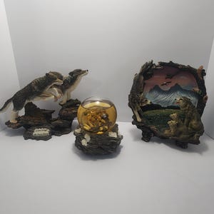 May include: Three decorative wolf figurines and a snow globe. The figurines depict wolves in various poses, including a pair on a rocky base and a wolf family scene. The snow globe features a wolf and small objects.
