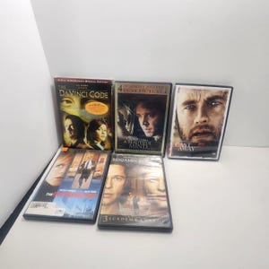 May include: A collection of five DVD cases featuring movie titles including The Da Vinci Code, A Beautiful Mind, Cast Away, The Interpreter, and The Curious Case of Benjamin Button, each with distinct cover art displaying actors and film information.