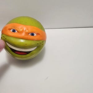 Könnte beinhalten: Ein grünes, kugelförmiges Spielzeug mit dem Gesicht von Michelangelo von den Teenage Mutant Ninja Turtles. Das Spielzeug hat eine orangefarbene Maske, blaue Augen und einen lächelnden Mund. Es ist ein Sammlerstück.