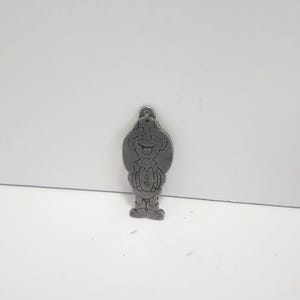 May include: A silver-colored metal charm in the shape of a cartoon character. The charm features a smiling face, a hat, and a button-down vest. The charm has a small loop at the top for attaching to a keychain or necklace.