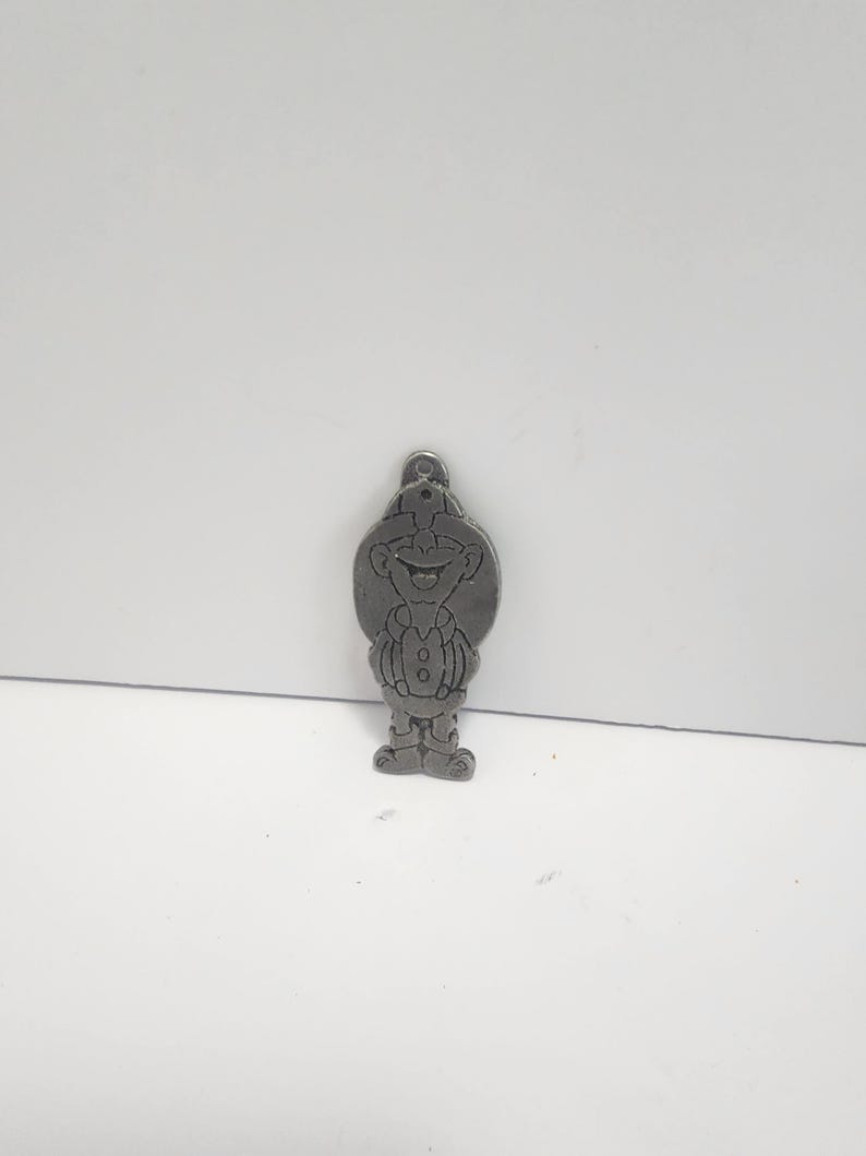 May include: A silver-colored metal charm in the shape of a cartoon character. The charm features a smiling face, a hat, and a button-down vest. The charm has a small loop at the top for attaching to a keychain or necklace.
