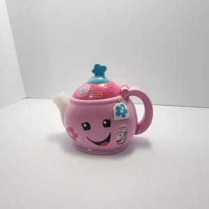 May include: A pink Fisher-Price Smart Stages teapot toy with a smiling face, flowers, and a blue lid. The teapot has a handle and a spout. The toy has the words "Smart Stages" printed on the side.