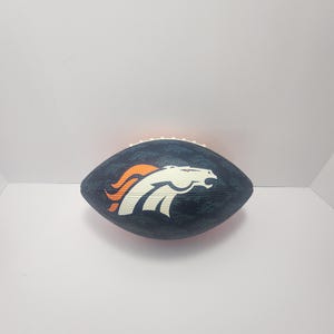 Könnte beinhalten: Ein marineblauer Football mit dem Logo der Denver Broncos in Weiß, Orange und Marineblau. Der Football ist oval und hat weiße Nähte an den Rändern.