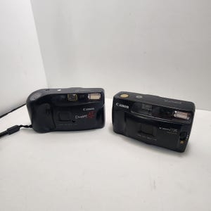 May include: Two black Canon point-and-shoot film cameras. The left camera is a Canon Snappy AF with a wrist strap. The right camera is a Canon Snappy TX Macro. Both cameras feature a built-in flash, a lens, and a viewfinder on the front body.